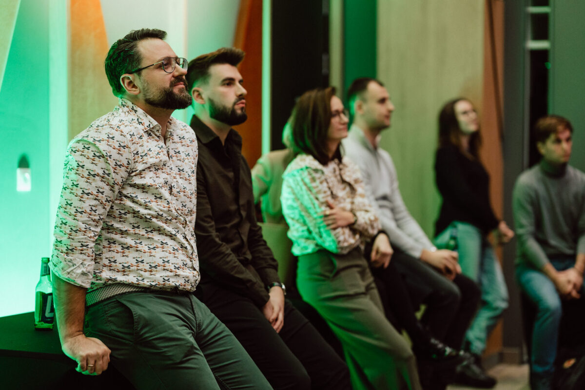 zdjecia eventowe aula polska bnp paribas warszawa 027 Grupa sześciu osób siedzi i stoi pod ścianą w nowoczesnym, delikatnie oświetlonym pomieszczeniu, uważnie obserwując coś poza kamerą. Atmosfera wydaje się spokojna i skupiona.