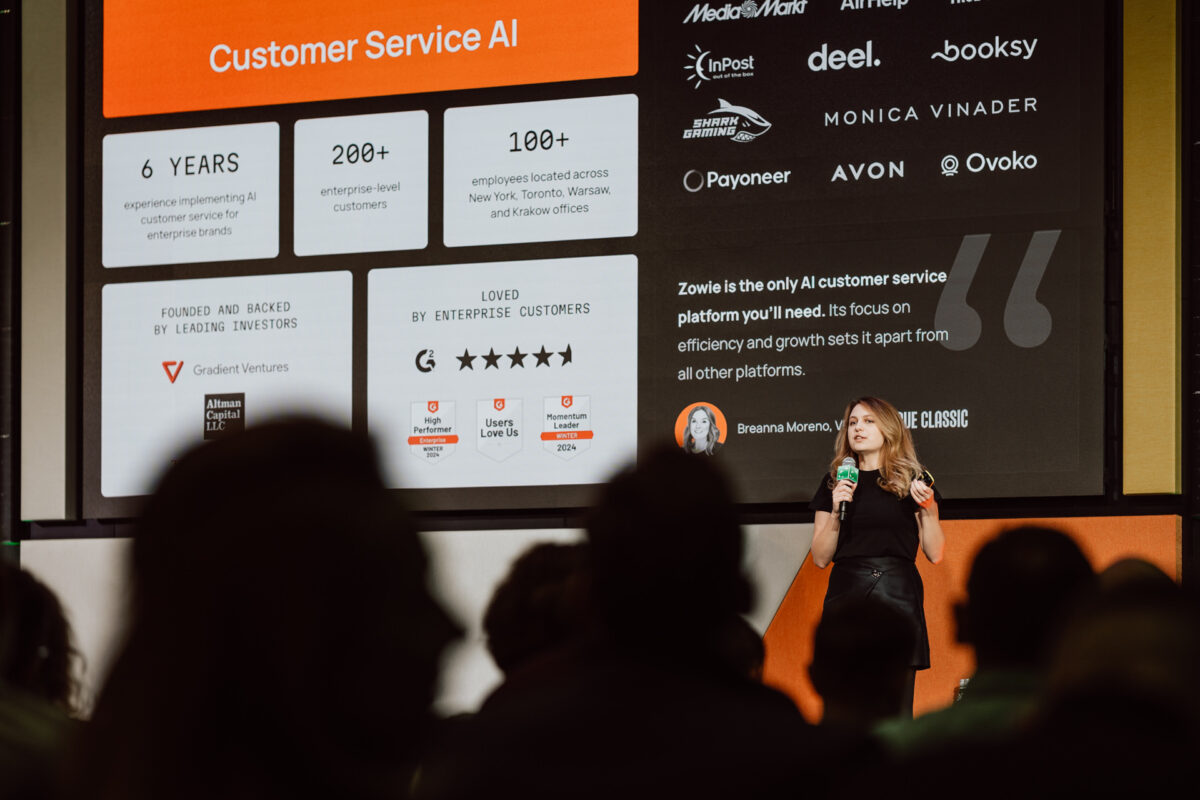 zdjecia eventowe aula polska bnp paribas warszawa 028 Kobieta stoi na scenie, prowadząc prezentację na temat sztucznej inteligencji w obsłudze klienta. Za nią znajduje się duży ekran wyświetlający statystyki firmy, logo, referencje i funkcje, podczas gdy publiczność patrzy.
