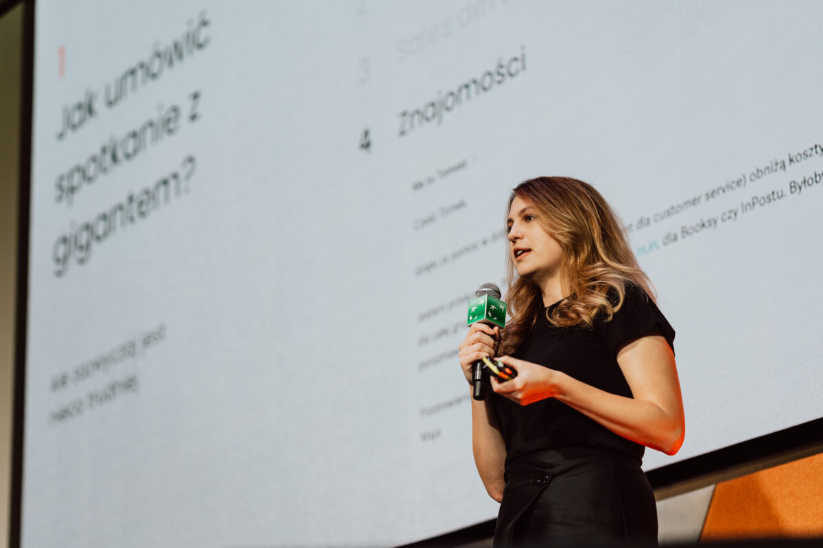 zdjecia eventowe aula polska bnp paribas warszawa 031 Kobieta trzymająca mikrofon i pilot do prezentacji przemawia na scenie przed dużym ekranem wyświetlającym tekst w języku polskim.