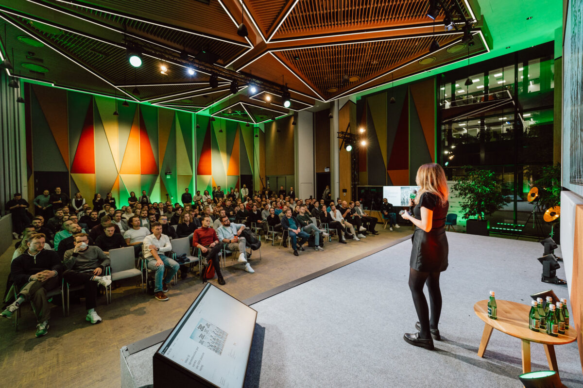 zdjecia eventowe aula polska bnp paribas warszawa 032 Kobieta stoi na scenie, przemawiając do dużej publiczności siedzącej w nowoczesnej, jasno oświetlonej sali konferencyjnej z kolorowymi geometrycznymi panelami ściennymi. Na ekranie obok niej widoczny jest slajd prezentacji.
