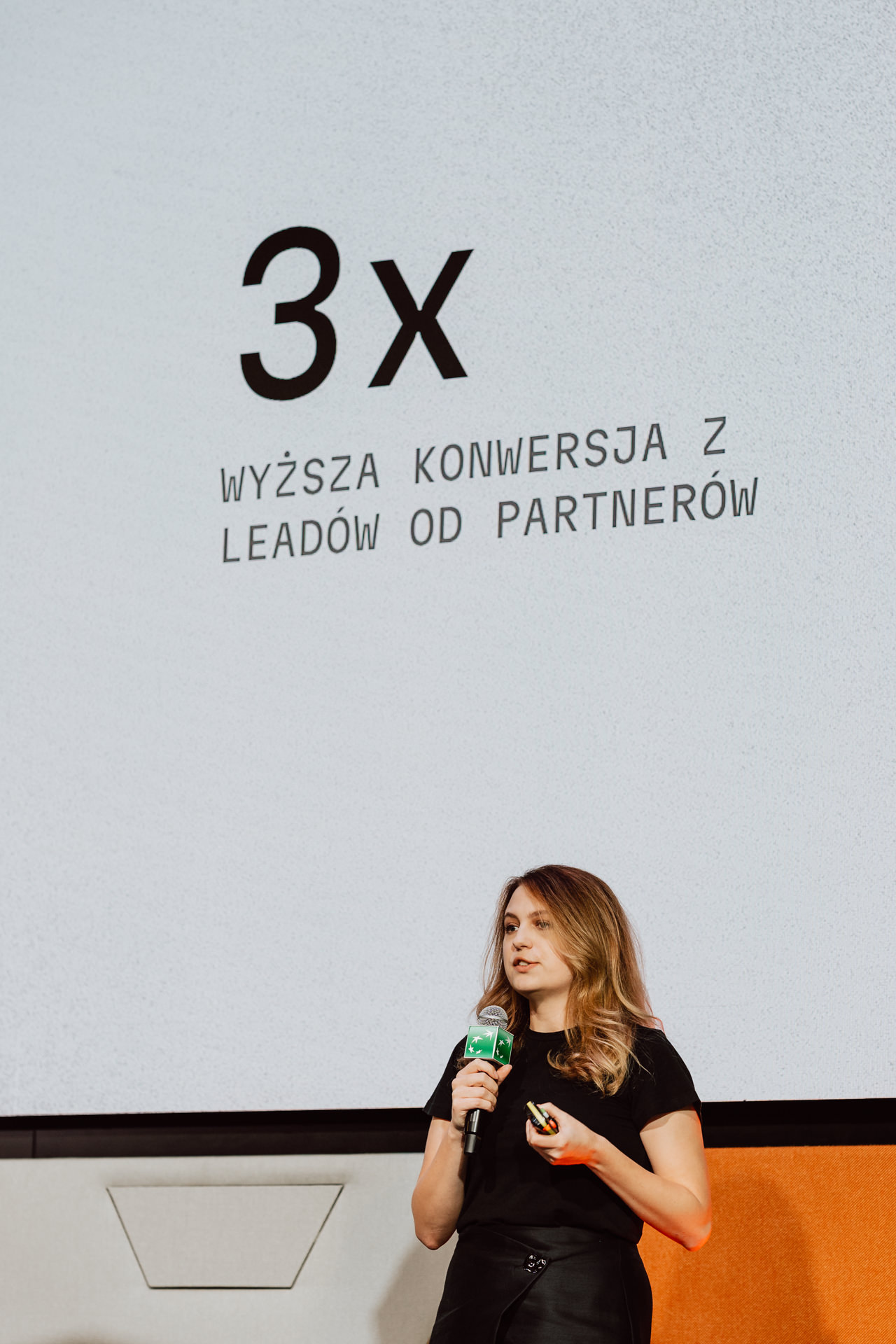 Kobieta stoi i mówi do mikrofonu przed slajdem prezentacji z napisem "3x WYŻSZA KONWERSJA Z LEADÓW OD PARTNERÓW" w języku polskim.