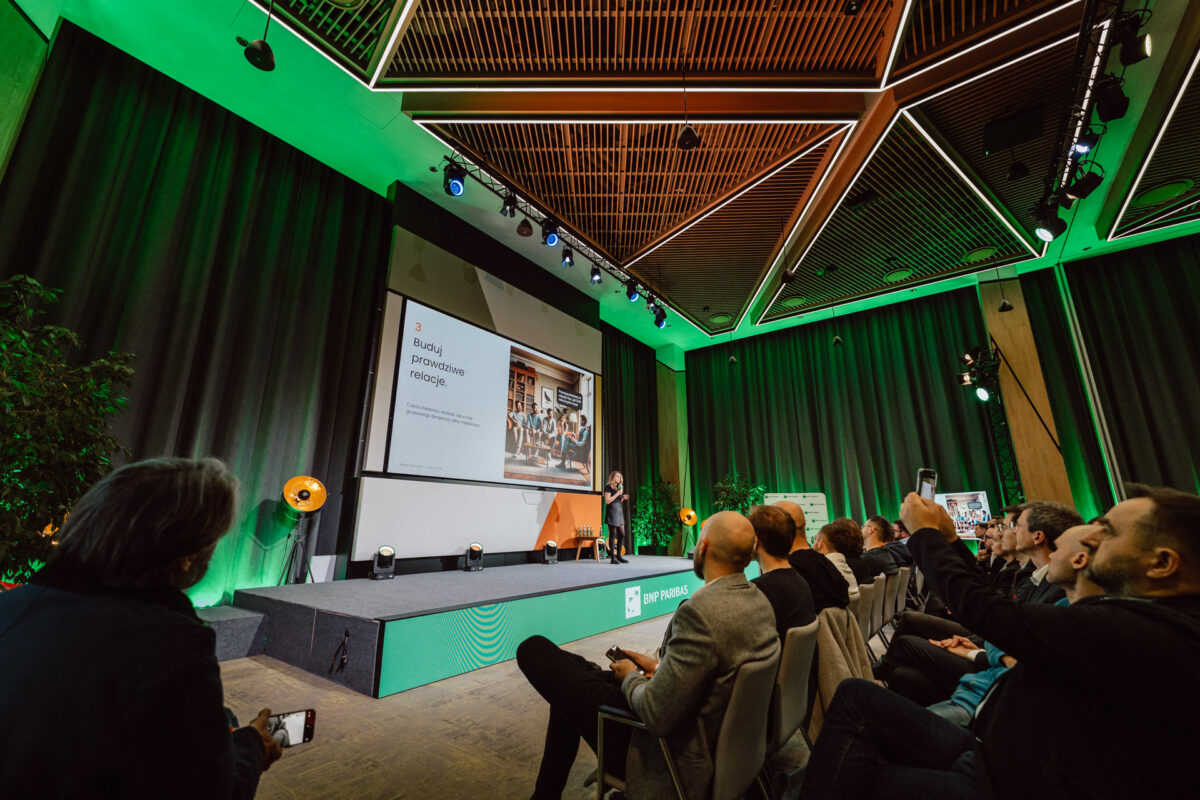 zdjecia eventowe aula polska bnp paribas warszawa 037 Prelegent stoi na scenie, prezentując publiczności w nowoczesnej, dobrze oświetlonej sali konferencyjnej z zielonym oświetleniem i dużym ekranem wyświetlającym slajd. Ludzie na widowni oglądają i robią zdjęcia.