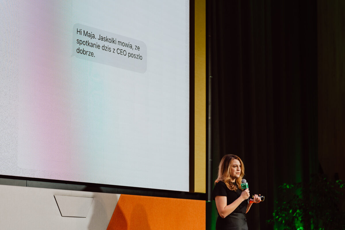 zdjecia eventowe aula polska bnp paribas warszawa 038 Kobieta trzymająca mikrofon stoi na scenie w pobliżu dużego ekranu wyświetlającego komunikat w języku polskim, który brzmi: "Cześć Maja. Jaskółki mówią, że spotkanie dziś z CEO poszło dobrze.