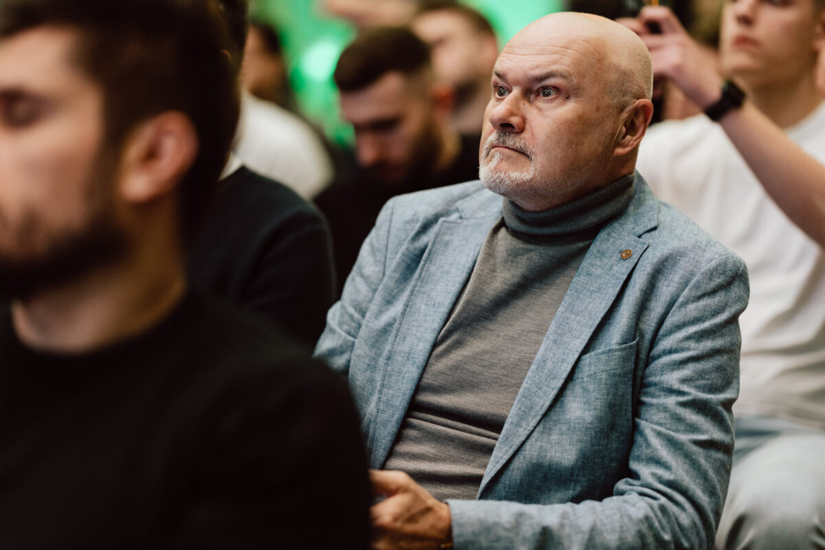 zdjecia eventowe aula polska bnp paribas warszawa 039 Łysy mężczyzna z siwą brodą, ubrany w jasnoniebieską marynarkę i szary golf, siedzi uważnie na widowni wśród innych osób, z których niektóre są nieostre na pierwszym planie i w tle.