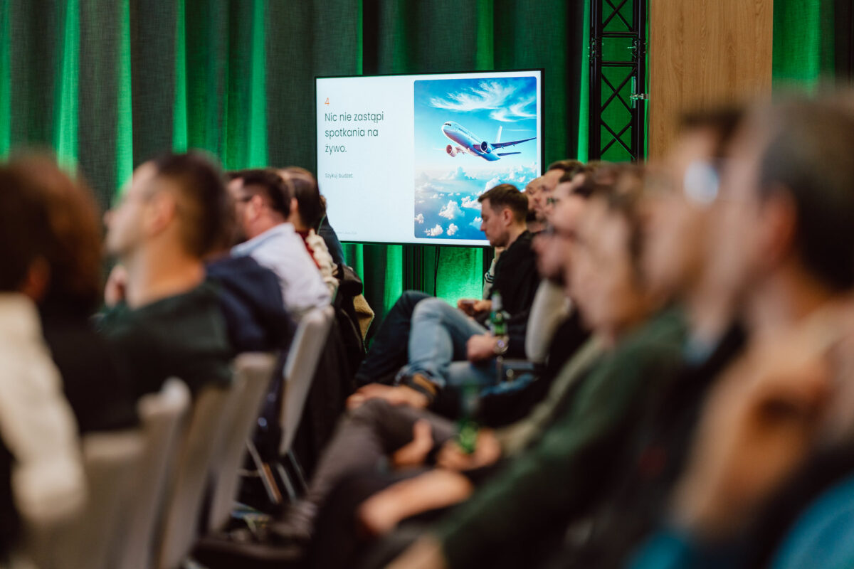 zdjecia eventowe aula polska bnp paribas warszawa 040 Grupa ludzi siedzi w rzędach, słuchając prezentacji. Na ekranie wyświetlany jest obraz samolotu na niebie i polski tekst, a w tle kurtyna i drewniany panel. Atmosfera jest skupiona i uważna.