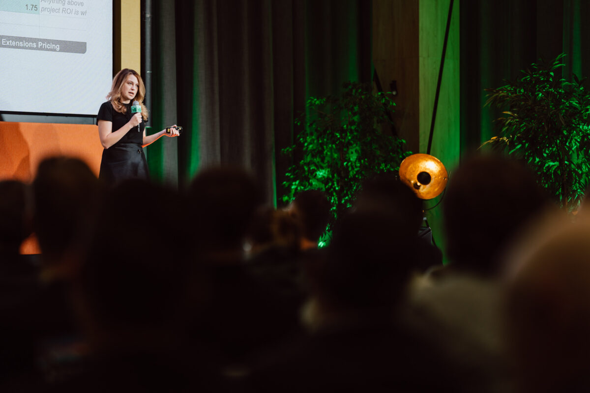 zdjecia eventowe aula polska bnp paribas warszawa 041 Kobieta stoi na scenie, trzymając mikrofon i przemawiając do publiczności. Znajduje się w pobliżu ekranu wyświetlającego prezentację, z zielonymi roślinami i dużym reflektorem w tle.