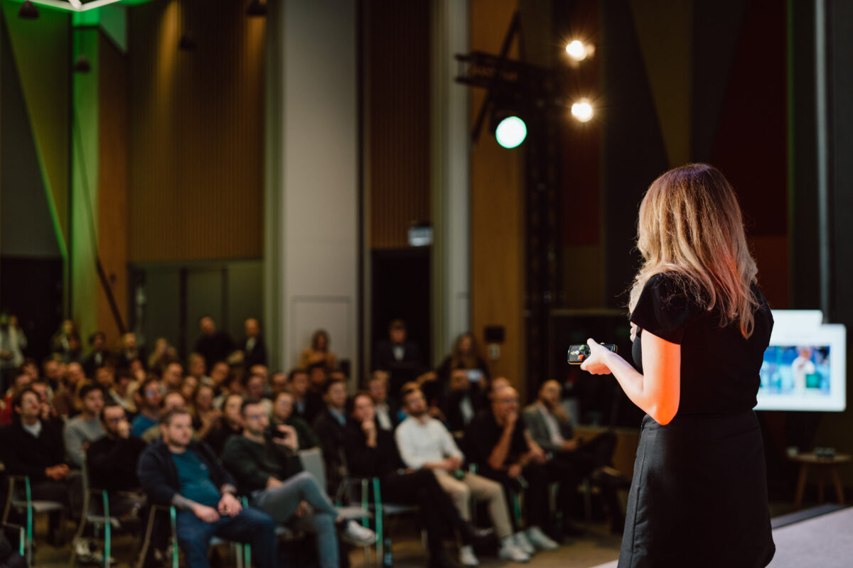 zdjecia eventowe aula polska bnp paribas warszawa 042 Kobieta z długimi włosami stoi na scenie, trzymając mikrofon i twarzą do siedzącej publiczności w nowoczesnej sali konferencyjnej. Publiczność słucha uważnie. Jasne światła oświetlają przestrzeń.
