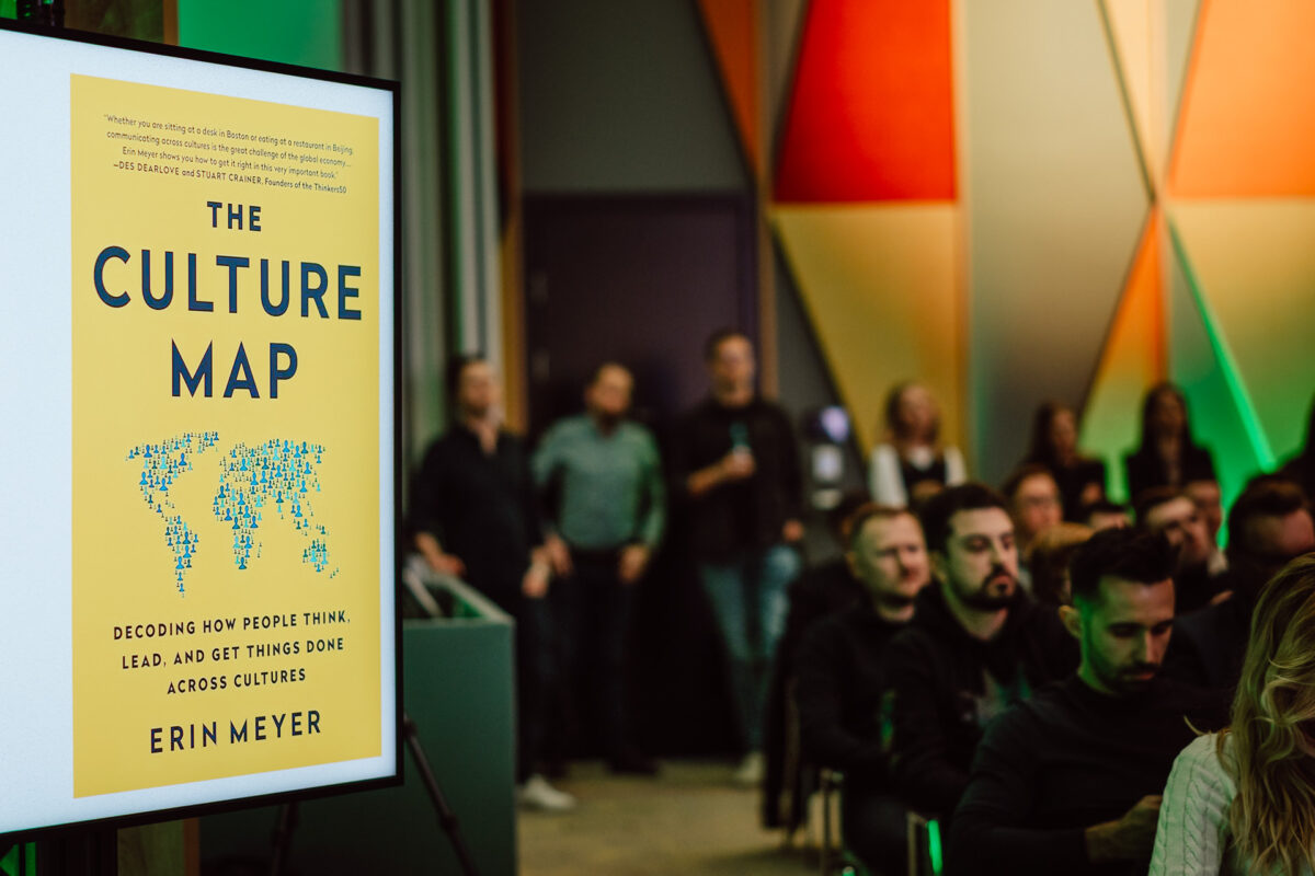 zdjecia eventowe aula polska bnp paribas warszawa 043 W nowoczesnym pomieszczeniu siedzi publiczność prowadząca prezentację. Na pierwszym planie duży ekran wyświetla okładkę książki "The Culture Map" autorstwa Erin Meyer, z żółtym tłem i mapą świata.