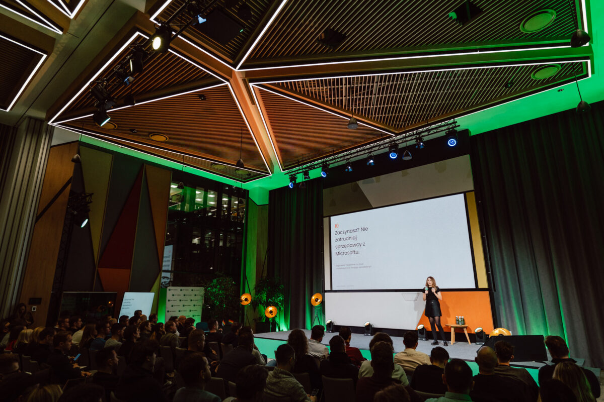 zdjecia eventowe aula polska bnp paribas warszawa 044 Kobieta stoi na scenie, prowadząc prezentację dla siedzącej publiczności w nowoczesnej sali konferencyjnej z geometrycznymi wzorami sufitowymi, jasnym oświetleniem scenicznym i dużym ekranem projektora wyświetlającym tekst.