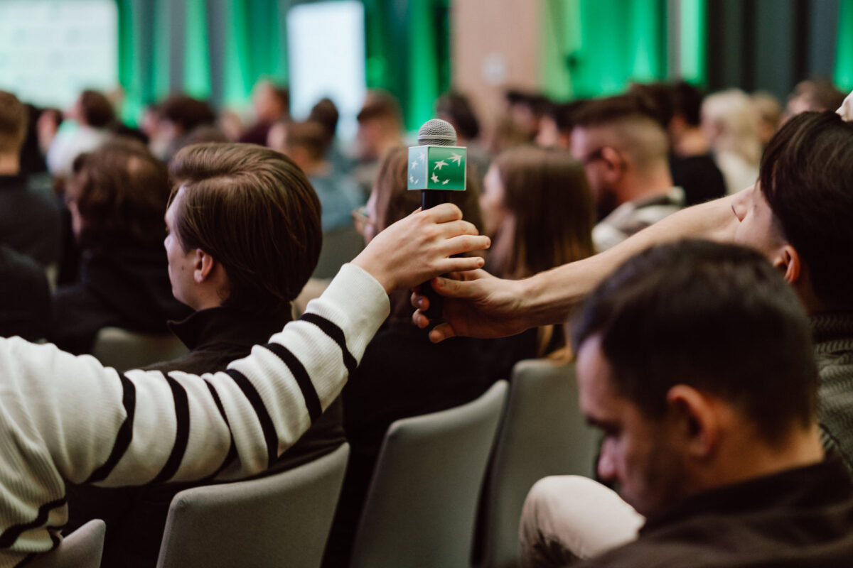 zdjecia eventowe aula polska bnp paribas warszawa 045 Osoba podaje mikrofon innemu uczestnikowi w zatłoczonej sali konferencyjnej. Publiczność siedzi twarzą do przodu, a tło jest podświetlone na zielono.