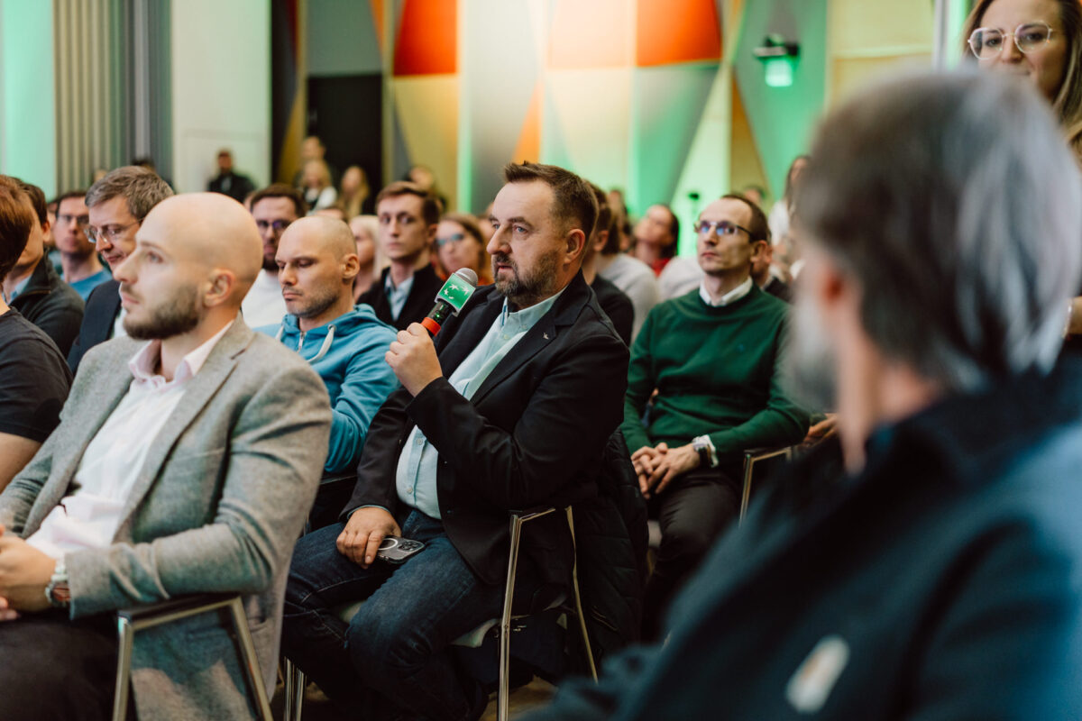 zdjecia eventowe aula polska bnp paribas warszawa 046 Mężczyzna trzymający mikrofon zadaje pytanie podczas konferencji lub seminarium, otoczony przez innych siedzących uczestników słuchających uważnie w nowoczesnym, dobrze oświetlonym pomieszczeniu.