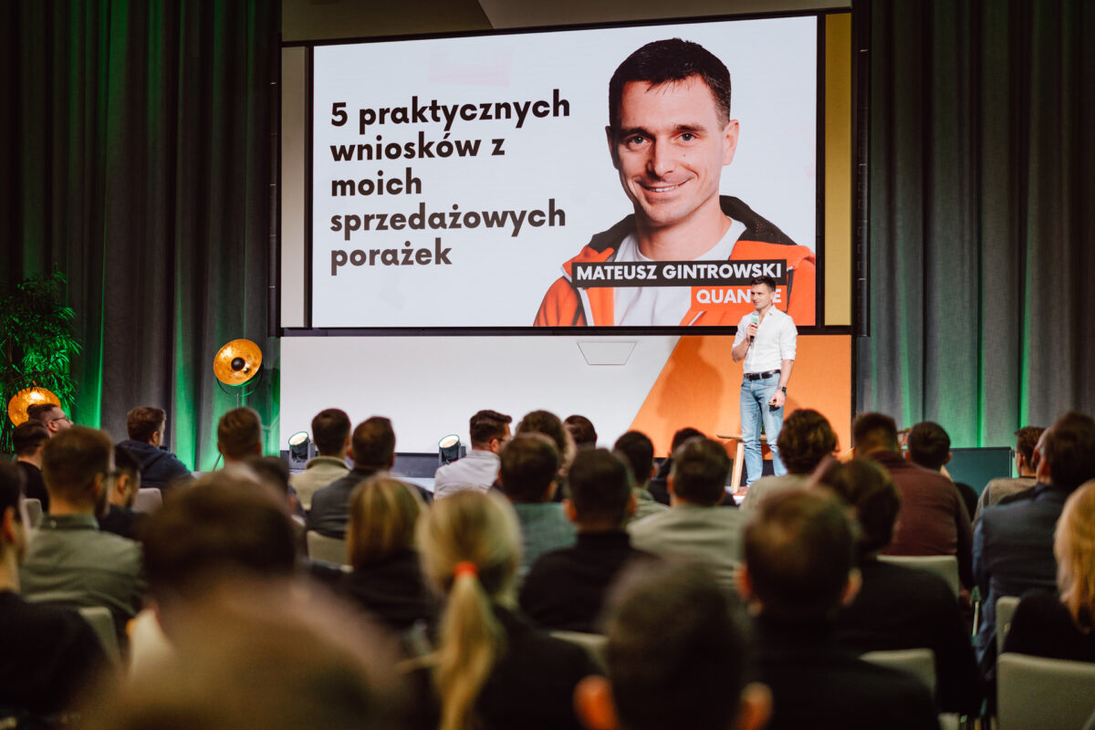 zdjecia eventowe aula polska bnp paribas warszawa 048 Mężczyzna stoi na scenie i przemawia do publiczności. Za nim znajduje się duży ekran wyświetlający jego zdjęcie i polski tekst: "5 praktycznych wniosków z moich sprzedażowych porażek" i "Mateusz Gintrowski.