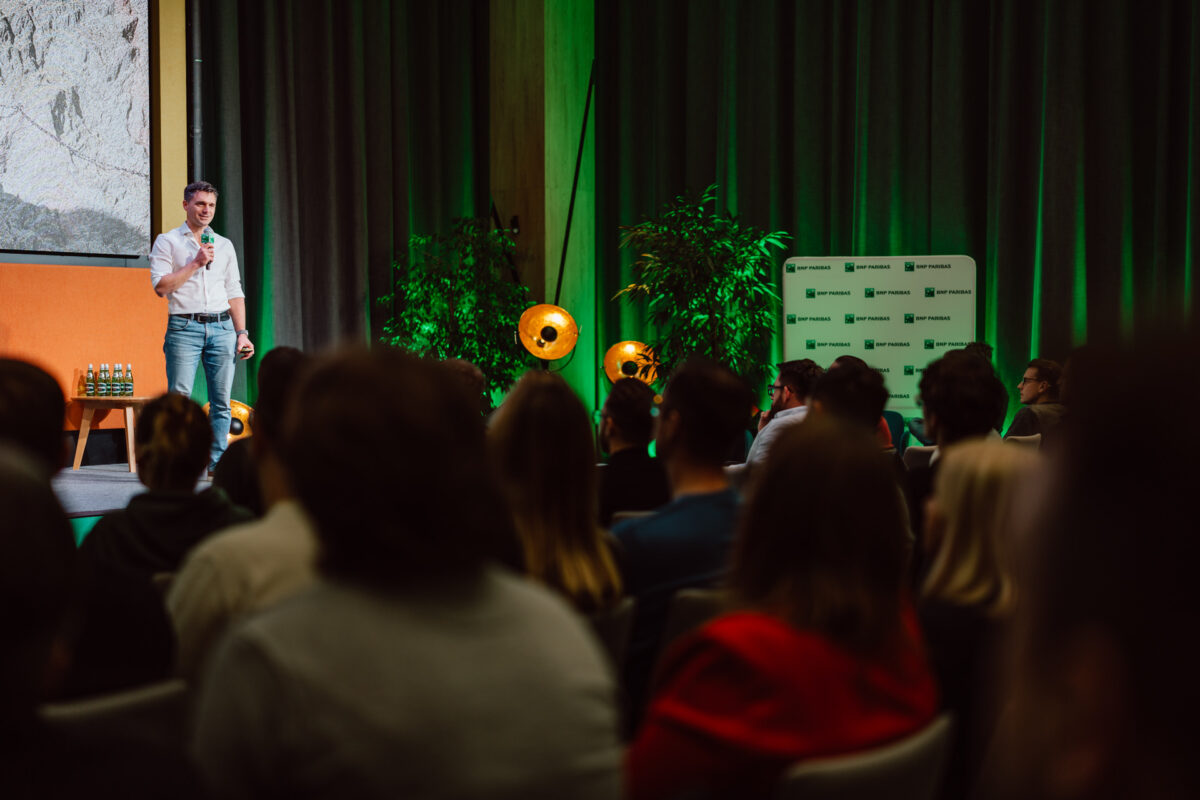 zdjecia eventowe aula polska bnp paribas warszawa 049 Mężczyzna w białej koszuli i dżinsach przemawia na scenie do publiczności w słabo oświetlonym pomieszczeniu z zielonym oświetleniem, roślinami i markowym tłem.