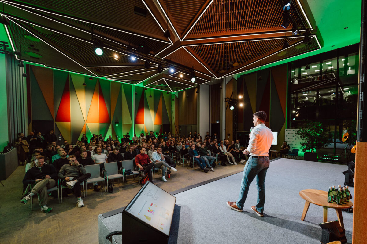 zdjecia eventowe aula polska bnp paribas warszawa 051 fotografia wydarzenia startupowego - Osoba stoi na scenie, prowadząc prezentację dla dużej publiczności w nowoczesnej, dobrze oświetlonej sali konferencyjnej z geometrycznymi wzorami na ścianach i zielonym oświetleniem akcentującym.