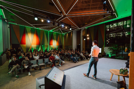 zdjecia eventowe aula polska bnp paribas warszawa 051 fotografia wydarzenia startupowego - Osoba stoi na scenie, prowadząc prezentację dla dużej publiczności w nowoczesnej, dobrze oświetlonej sali konferencyjnej z geometrycznymi wzorami na ścianach i zielonym oświetleniem akcentującym.