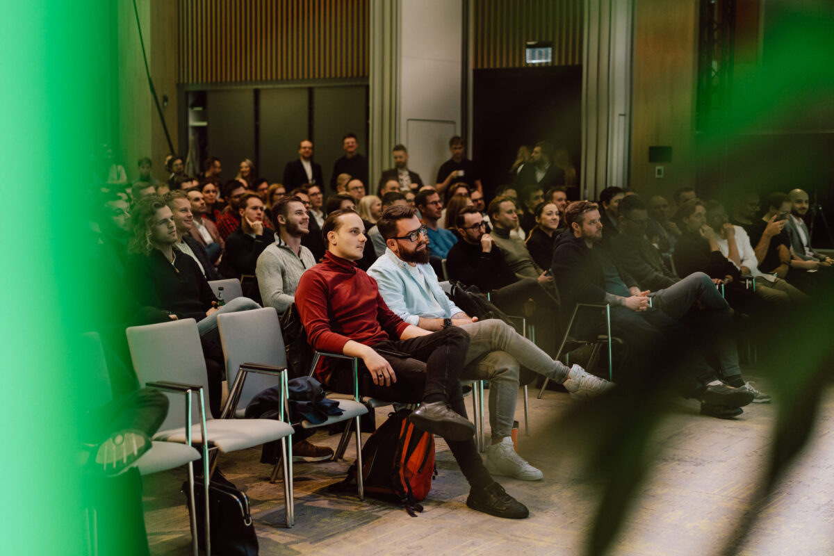 zdjecia eventowe aula polska bnp paribas warszawa 052 Grupa ludzi siedzi uważnie w rzędach krzeseł podczas imprezy lub konferencji w pomieszczeniu, a zieleń częściowo otacza pierwszy plan. Większość uczestników wydaje się być zaangażowana i skupiona na prezentacji.