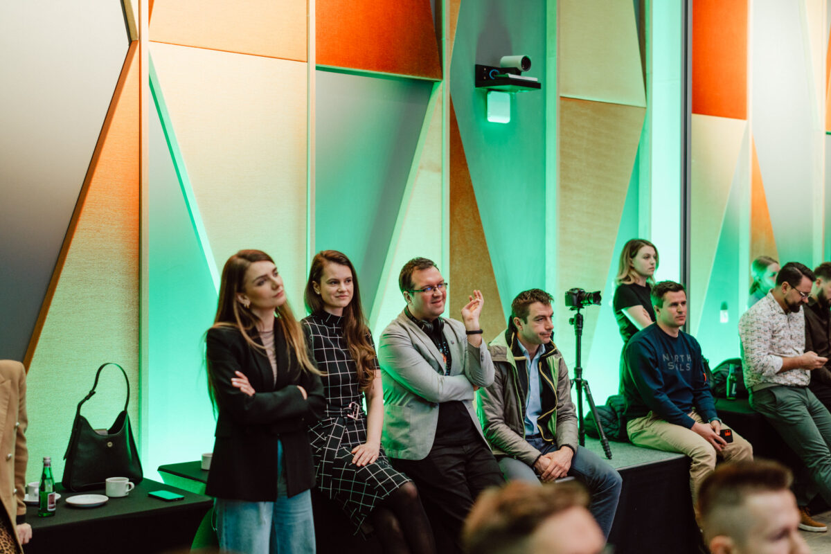 zdjecia eventowe aula polska bnp paribas warszawa 054 Grupa ludzi siedzi i stoi wzdłuż ściany z kolorowymi geometrycznymi panelami, obserwując wydarzenie. Niektórzy wyglądają na uważnych, podczas gdy inni wydają się zrelaksowani. Obok nich widoczna jest kamera i torby.