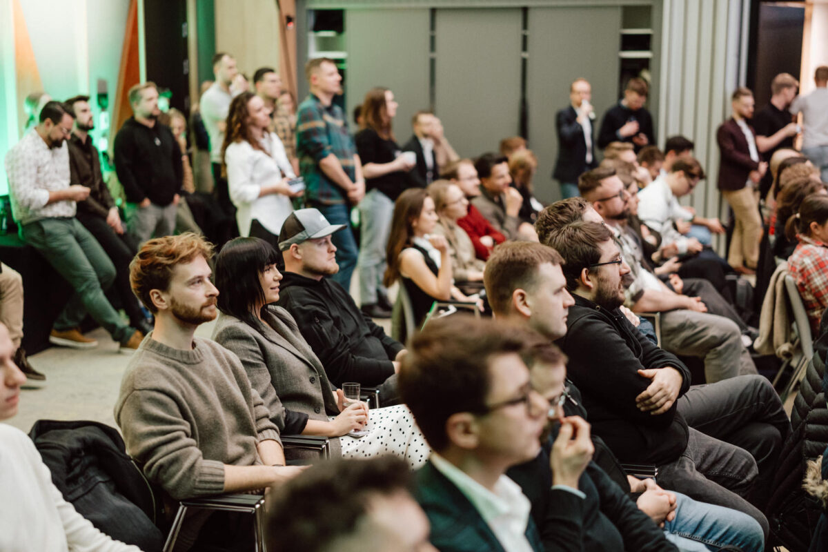 zdjecia eventowe aula polska bnp paribas warszawa 055 Zróżnicowana grupa ludzi siedzi i stoi uważnie w nowoczesnej sali konferencyjnej, oglądając prezentację lub prelegenta. Niektórzy wydają się być zaangażowani, podczas gdy inni swobodnie obserwują lub cicho rozmawiają.