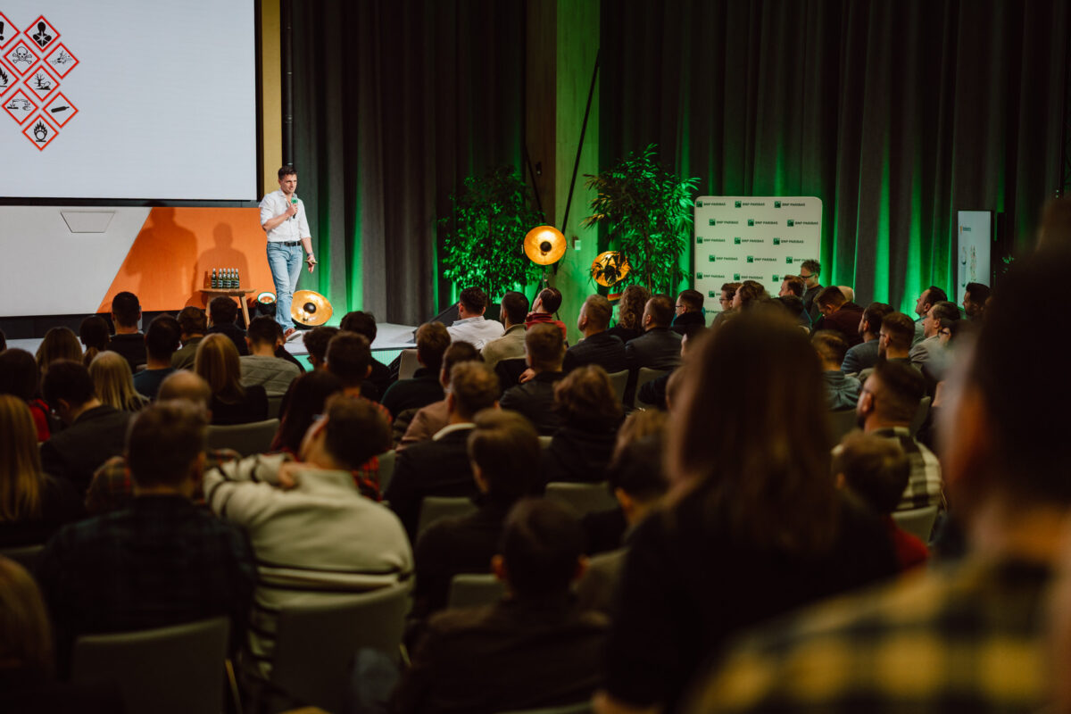 zdjecia eventowe aula polska bnp paribas warszawa 056 Mężczyzna stoi na scenie, prowadząc prezentację dla siedzącej publiczności w sali konferencyjnej, z dużym ekranem, zielonymi zasłonami i ciepłym oświetleniem w tle.