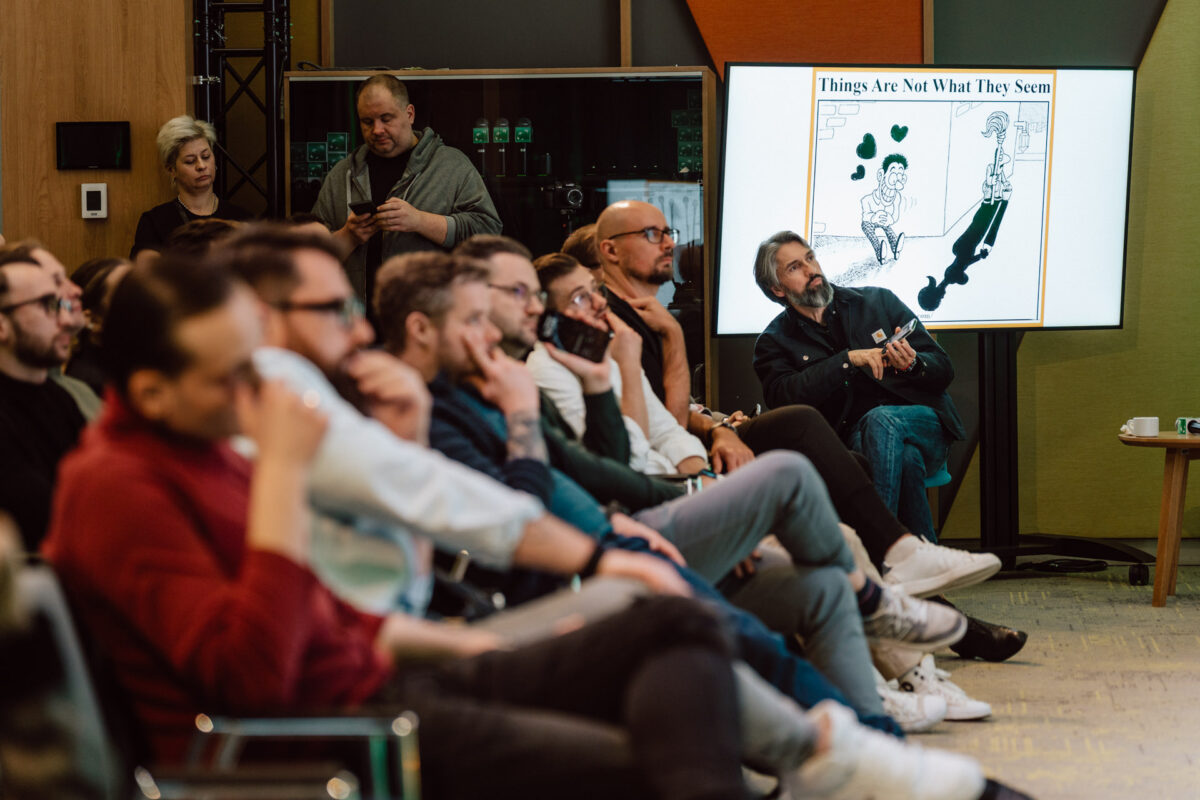 zdjecia eventowe aula polska bnp paribas warszawa 062 Grupa ludzi siedzi i słucha uważnie w nowoczesnym pokoju, podczas gdy mężczyzna gestykuluje w kierunku ekranu wyświetlającego kreskówkę zatytułowaną "Rzeczy nie są tym, czym się wydają".