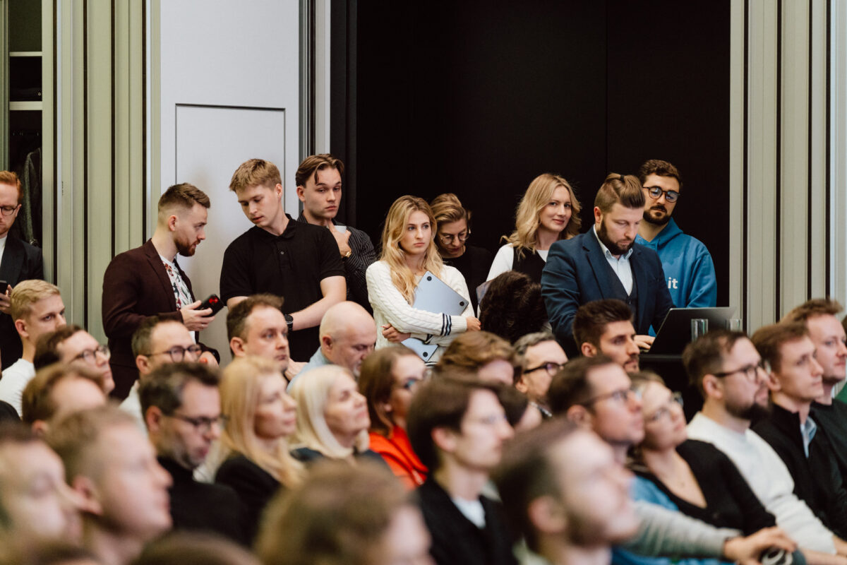 zdjecia eventowe aula polska bnp paribas warszawa 063 Grupa ludzi stoi z tyłu zatłoczonego pomieszczenia podczas oficjalnego wydarzenia. Większość z nich uważnie obserwuje, niektórzy trzymają laptopy lub telefony, podczas gdy wielu innych siedzi i skupia się na mówcy poza kadrem.