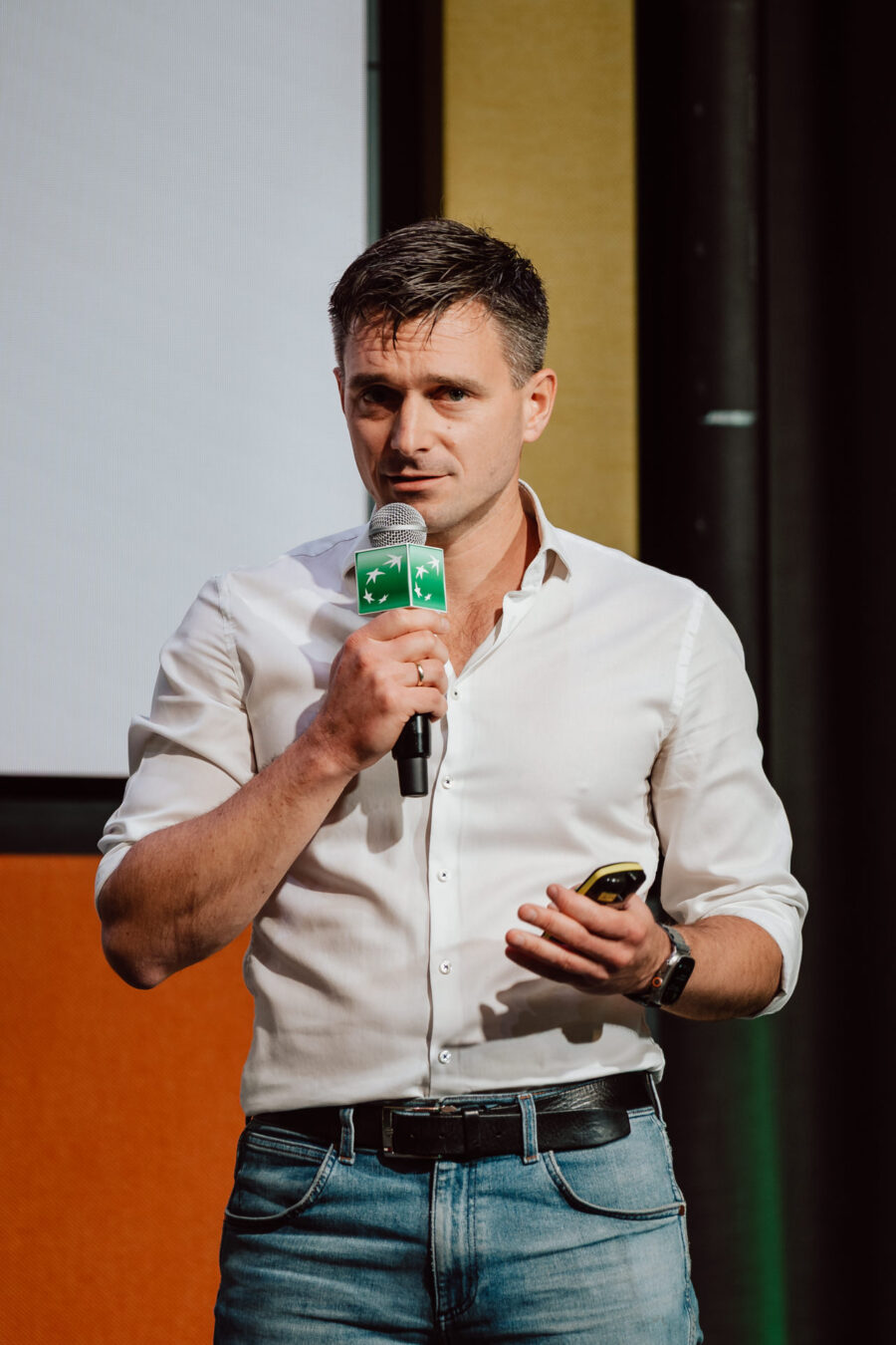 zdjecia eventowe aula polska bnp paribas warszawa 065 Mężczyzna w białej koszuli i dżinsach trzyma mikrofon i urządzenie podczas przemawiania na scenie. Stoi przed ekranem i pomarańczową ścianą, patrząc bezpośrednio na publiczność.