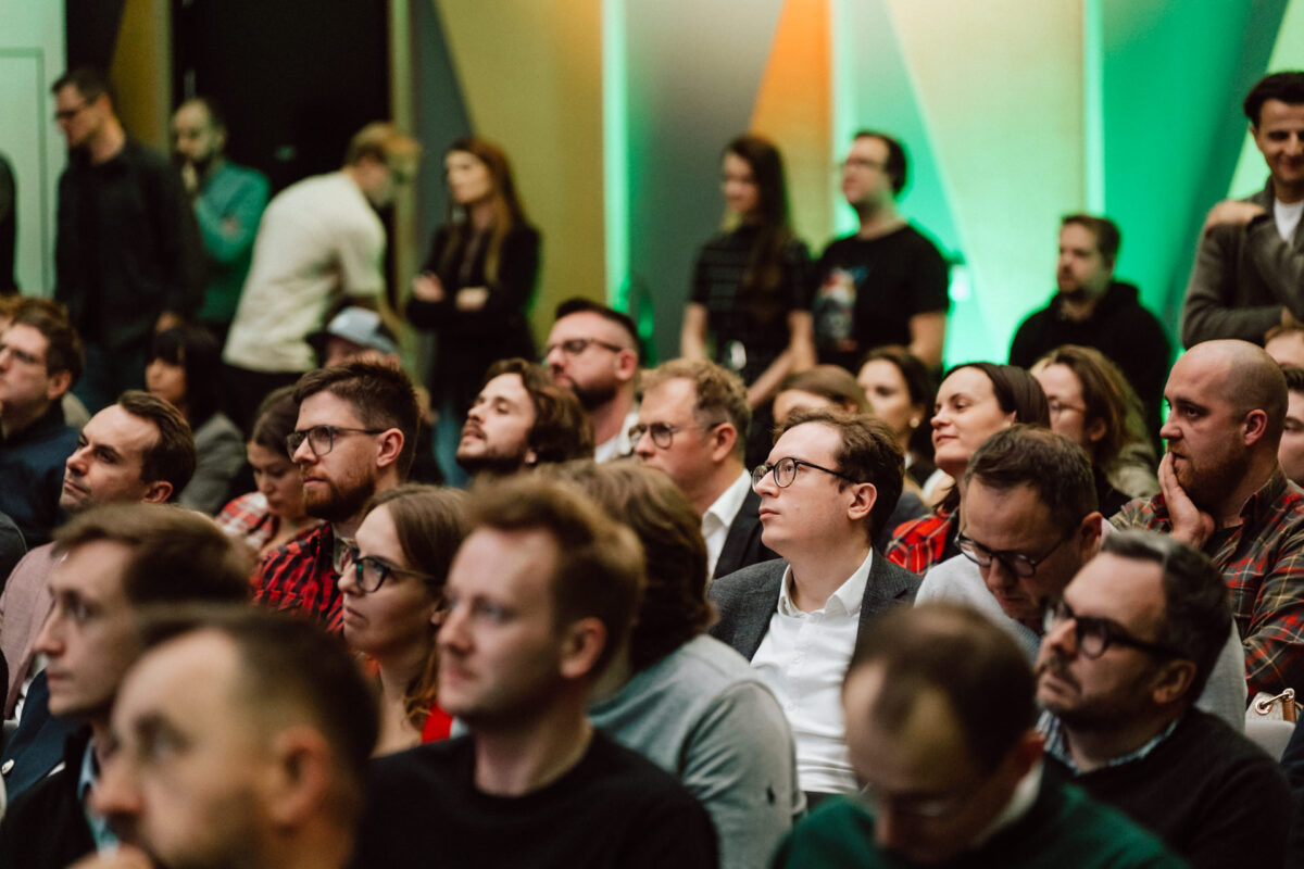 zdjecia eventowe aula polska bnp paribas warszawa 067 Grupa ludzi siedzi blisko siebie na widowni, uważnie słuchając wydarzenia lub prezentacji. Tło jest kolorowe, a niektóre osoby stoją. Większość uczestników wydaje się skupiona i zaangażowana.