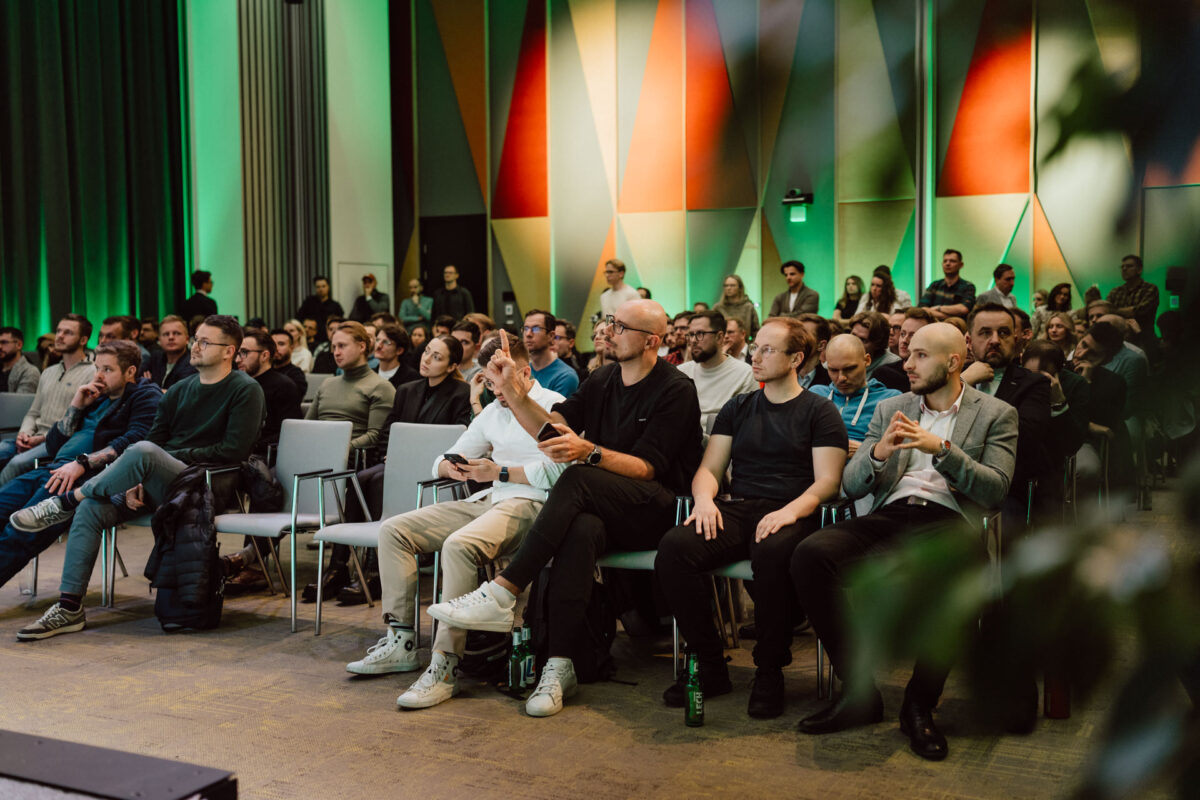 zdjecia eventowe aula polska bnp paribas warszawa 068 Duża grupa ludzi, głównie mężczyzn, siedzi w rzędach krzeseł na imprezie lub konferencji, uważnie słuchając prezentacji. Tło stanowią kolorowe, geometryczne panele ścienne.