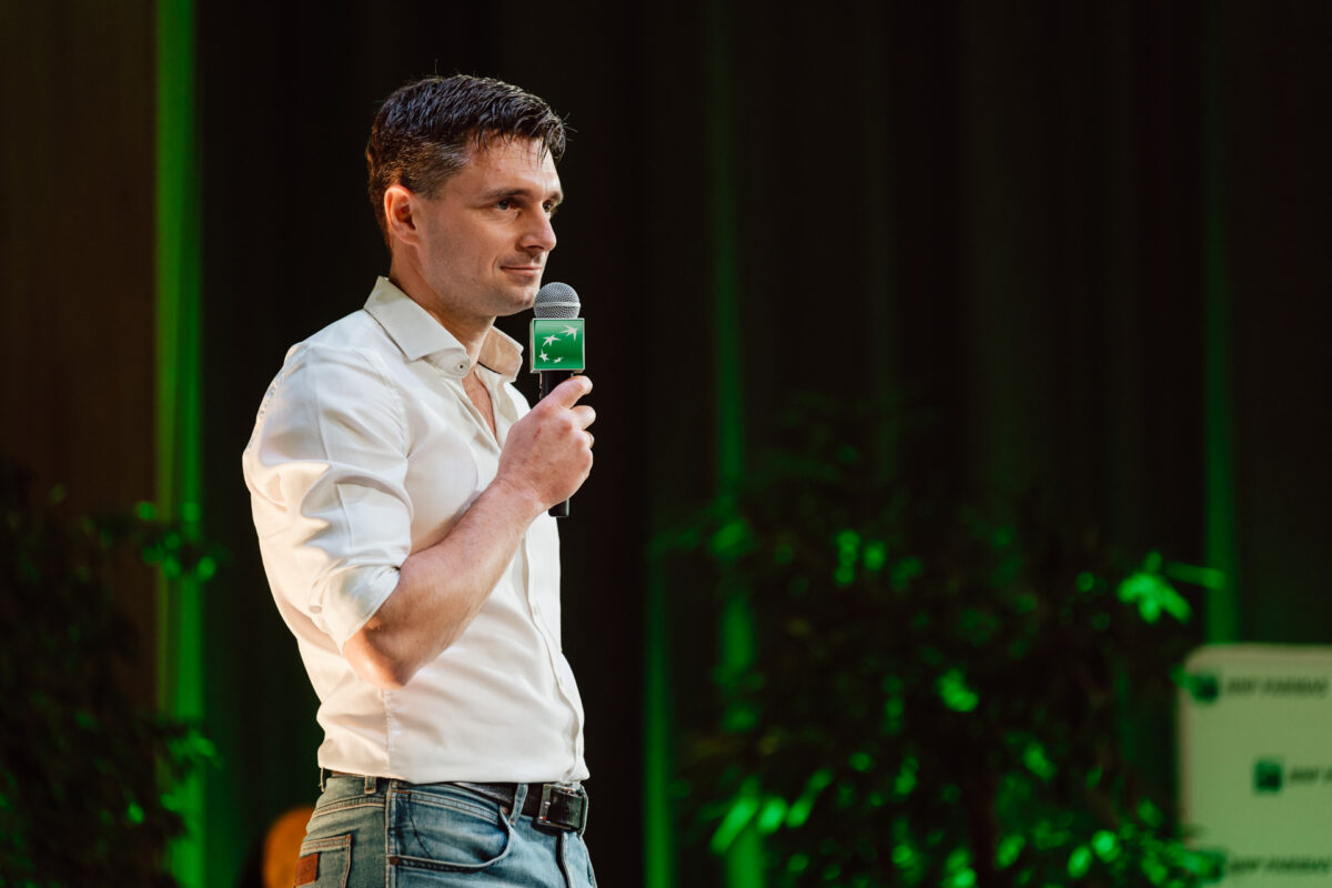zdjecia eventowe aula polska bnp paribas warszawa 069 Mężczyzna w białej koszuli i niebieskich dżinsach trzyma mikrofon i przemawia na scenie. Tło jest zielone z rozmytymi roślinami i częściowym widokiem na znak.