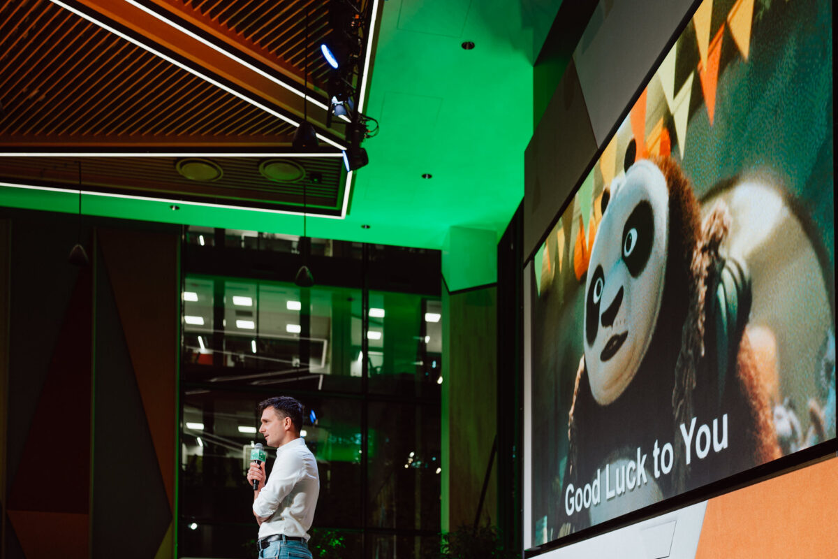 zdjecia eventowe aula polska bnp paribas warszawa 070 Mężczyzna trzymający mikrofon stoi na scenie przed dużym ekranem wyświetlającym animowaną pandę i tekst "Good Luck to You". W tle widoczne są kolorowe światła i nowoczesna architektura wnętrz.