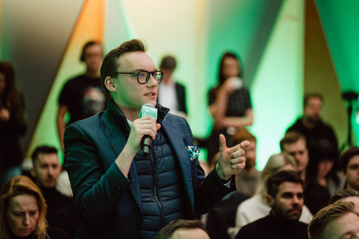 zdjecia eventowe aula polska bnp paribas warszawa 071 Osoba w okularach i niebieskiej marynarce mówi do mikrofonu w zatłoczonym pomieszczeniu, gestykulując ręką. Publiczność i zielone abstrakcyjne tło są widoczne za nimi.