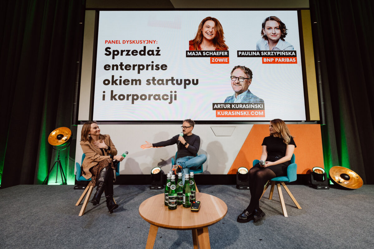 zdjecia eventowe aula polska bnp paribas warszawa 072 Trzech panelistów siedzi na scenie z mikrofonami w rękach, zaangażowanych w dyskusję. Duży ekran za nimi wyświetla ich imiona, zdjęcia i temat panelu w języku polskim, a butelki z wodą stoją na stole z przodu.