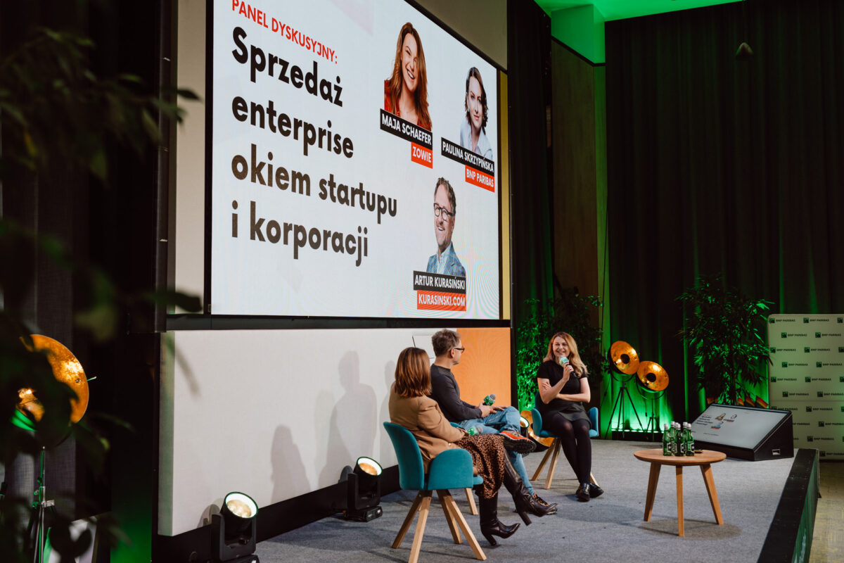 zdjecia eventowe aula polska bnp paribas warszawa 074 Trzy osoby siedzą na scenie i prowadzą dyskusję panelową. Za nimi duży ekran wyświetla temat w języku polskim: "Sprzedaż przedsiębiorstw okiem startupu i korporacji". Otoczenie jest nowoczesne, z zielonymi roślinami i ciepłym oświetleniem.