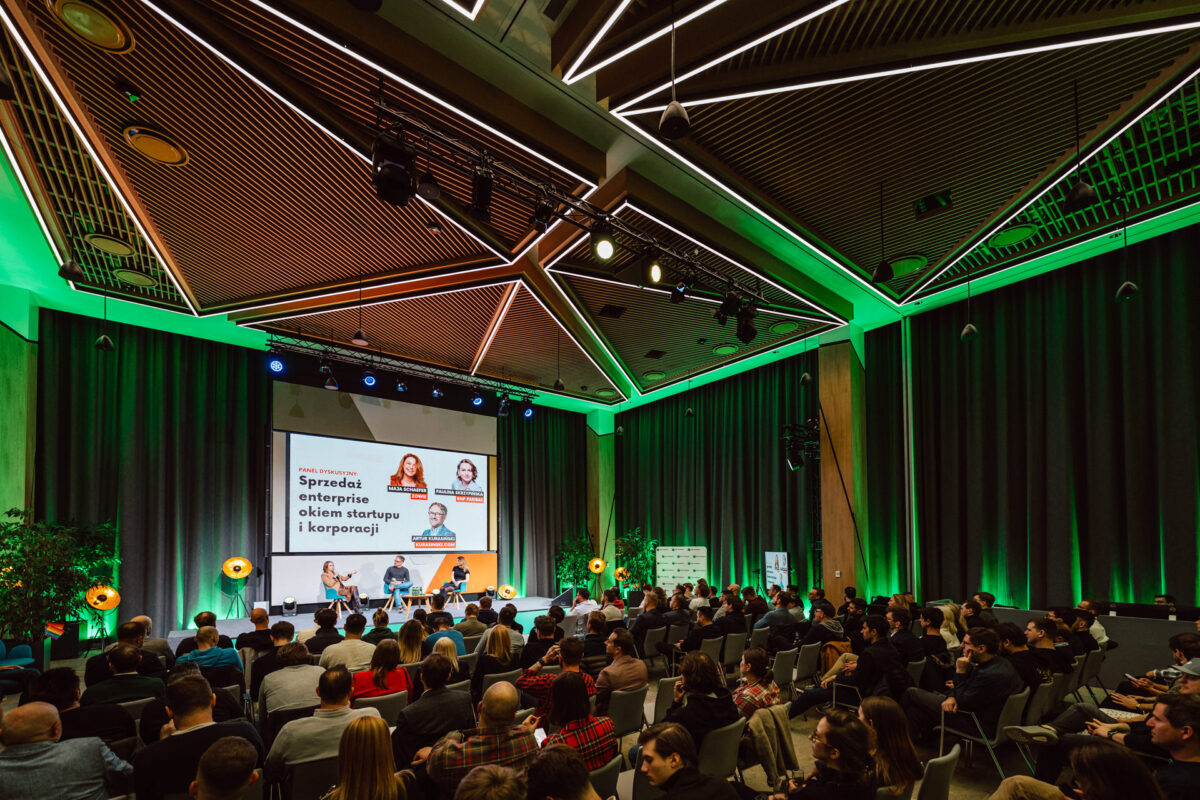 zdjecia eventowe aula polska bnp paribas warszawa 075 Sala konferencyjna wypełniona uczestnikami oglądającymi panel czterech prelegentów na scenie. Duży ekran za nimi wyświetla zdjęcia prelegentów i tekst w języku polskim. Pomieszczenie jest jasno oświetlone zielonym akcentem.