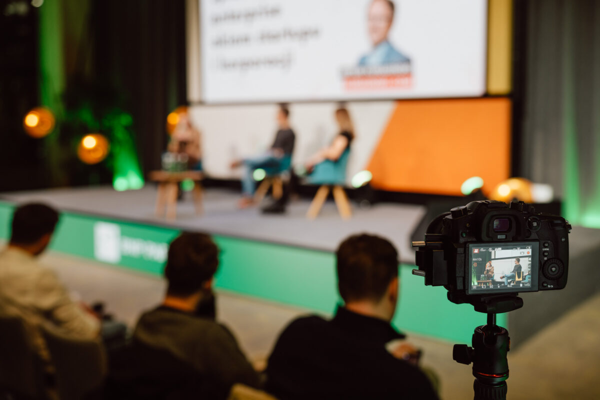 zdjecia eventowe aula polska bnp paribas warszawa 078 Kamera na statywie rejestruje dyskusję panelową na scenie, z dwoma niewyraźnymi prelegentami siedzącymi i publicznością na pierwszym planie. Scena jest jasno oświetlona, a w tle znajduje się duży ekran.