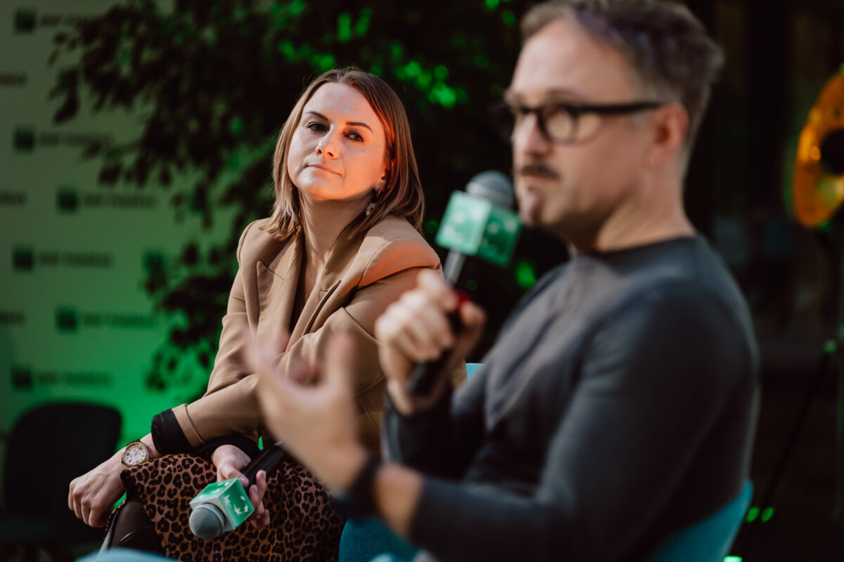 zdjecia eventowe aula polska bnp paribas warszawa 079 Kobieta w brązowej marynarce uważnie słucha, podczas gdy mężczyzna w okularach mówi do mikrofonu podczas imprezy w pomieszczeniu. Tło jest zielone z rozmytym brandingiem.