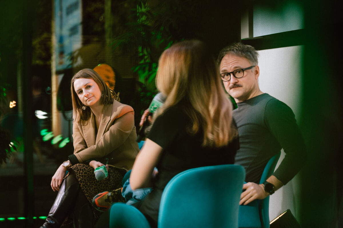 zdjecia eventowe aula polska bnp paribas warszawa 081 Trzy osoby siedzą na niebieskich krzesłach w nowoczesnym wnętrzu i prowadzą rozmowę. Kobieta w brązowej marynarce słucha uważnie, podczas gdy mężczyzna w okularach patrzy w stronę kamery. Trzecia osoba siedzi tyłem do kamery.