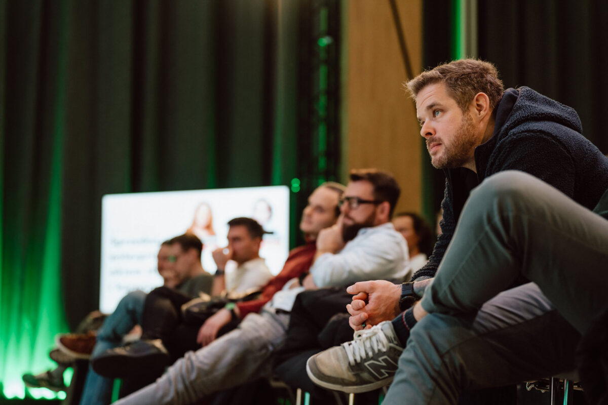 zdjecia eventowe aula polska bnp paribas warszawa 082 Grupa ludzi siedzi na krzesłach, uważnie oglądając coś poza kamerą. W tle znajduje się niewyraźny ekran prezentacji i ciemne zasłony, a zielone oświetlenie tworzy skupioną atmosferę.