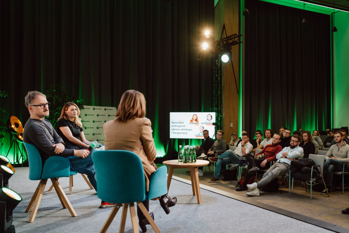 zdjecia eventowe aula polska bnp paribas warszawa 083 Trzy osoby siedzą na scenie naprzeciwko publiczności na imprezie wewnętrznej. Publiczność uważnie słucha. Na ekranie wyświetlana jest prezentacja ze zdjęciem i tekstem. Oświetlenie jest ciepłe z zielonymi akcentami.