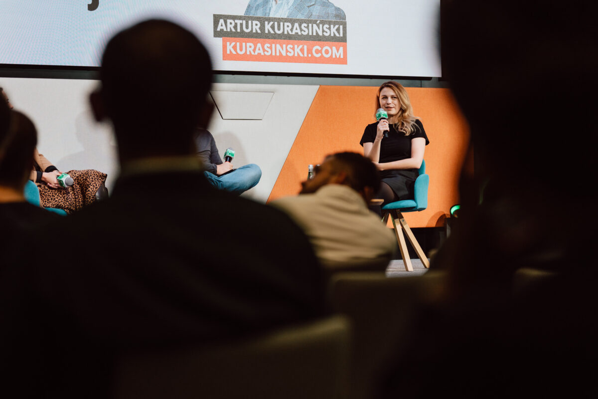 zdjecia eventowe aula polska bnp paribas warszawa 084 Kobieta przemawia na scenie z mikrofonem podczas panelu dyskusyjnego, siedząc na niebieskim krześle. Członkowie publiczności są widoczni na pierwszym planie. Powyżej widnieje napis "Artur Kurasiński.