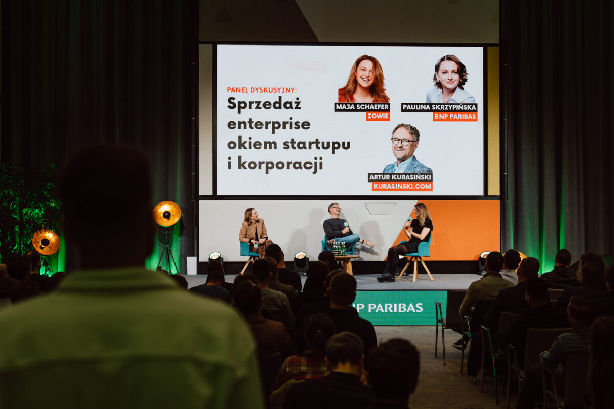 zdjecia eventowe aula polska bnp paribas warszawa 085 Panel dyskusyjny na scenie z trzema prelegentami siedzącymi, a za nimi ekran wyświetlający ich nazwiska, zdjęcia i temat napisany po polsku. Członkowie publiczności są widoczni na pierwszym planie.