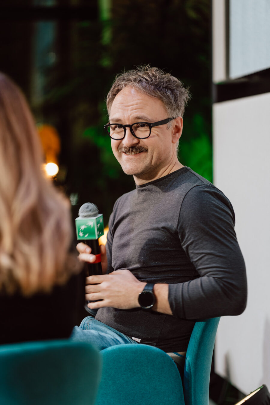 zdjecia eventowe aula polska bnp paribas warszawa 087 Mężczyzna w okularach i wąsach, ubrany w szarą koszulę i smartwatch, uśmiecha się, trzymając mikrofon podczas imprezy w pomieszczeniu. Siedzi na turkusowym krześle obok kobiety, której plecy zwrócone są w stronę kamery.