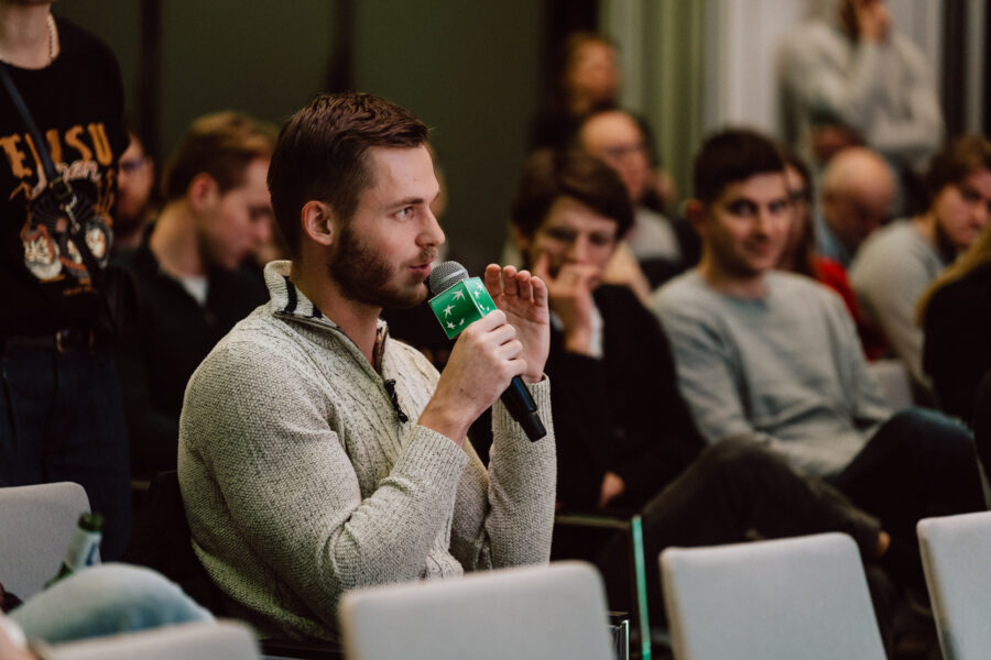 zdjecia eventowe aula polska bnp paribas warszawa 088 Mężczyzna w jasnym swetrze mówi do mikrofonu, siedząc wśród publiczności. Inni ludzie siedzą i słuchają uważnie w tle podczas czegoś, co wydaje się być konferencją lub seminarium.