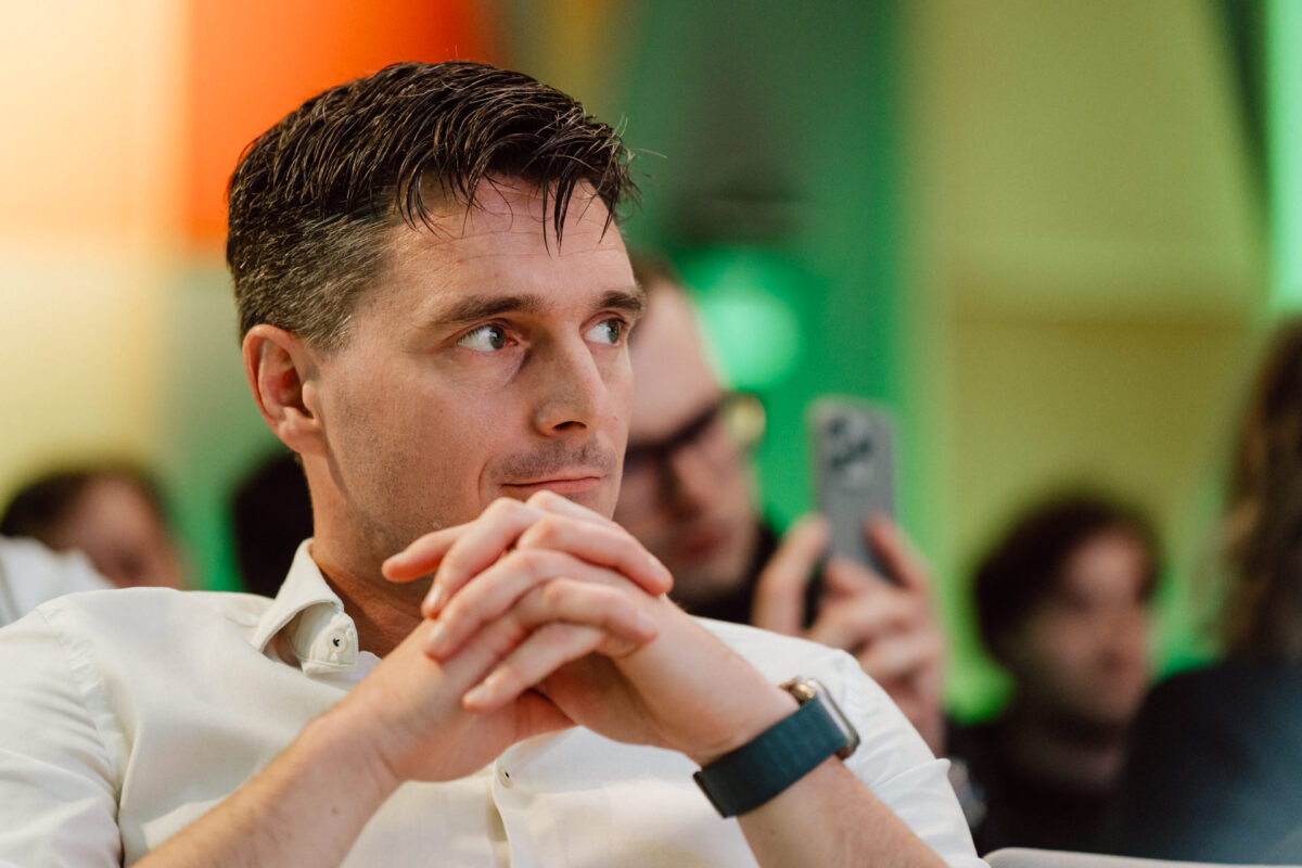 zdjecia eventowe aula polska bnp paribas warszawa 089 Mężczyzna z krótkimi ciemnymi włosami i w białej koszuli siedzi ze splecionymi dłońmi, patrząc w bok. Ludzie w tle są rozmazani, jeden trzyma smartfon. Oświetlenie jest kolorowe i miękkie.