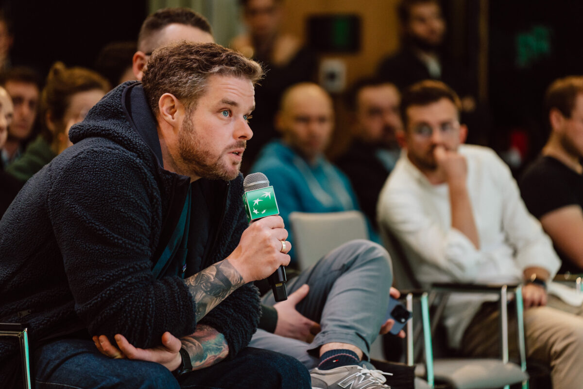 zdjecia eventowe aula polska bnp paribas warszawa 090 Mężczyzna z brodą i tatuażami trzyma mikrofon i przemawia, siedząc w grupie na imprezie w pomieszczeniu. Inni uczestnicy w swobodnych strojach uważnie słuchają w tle.