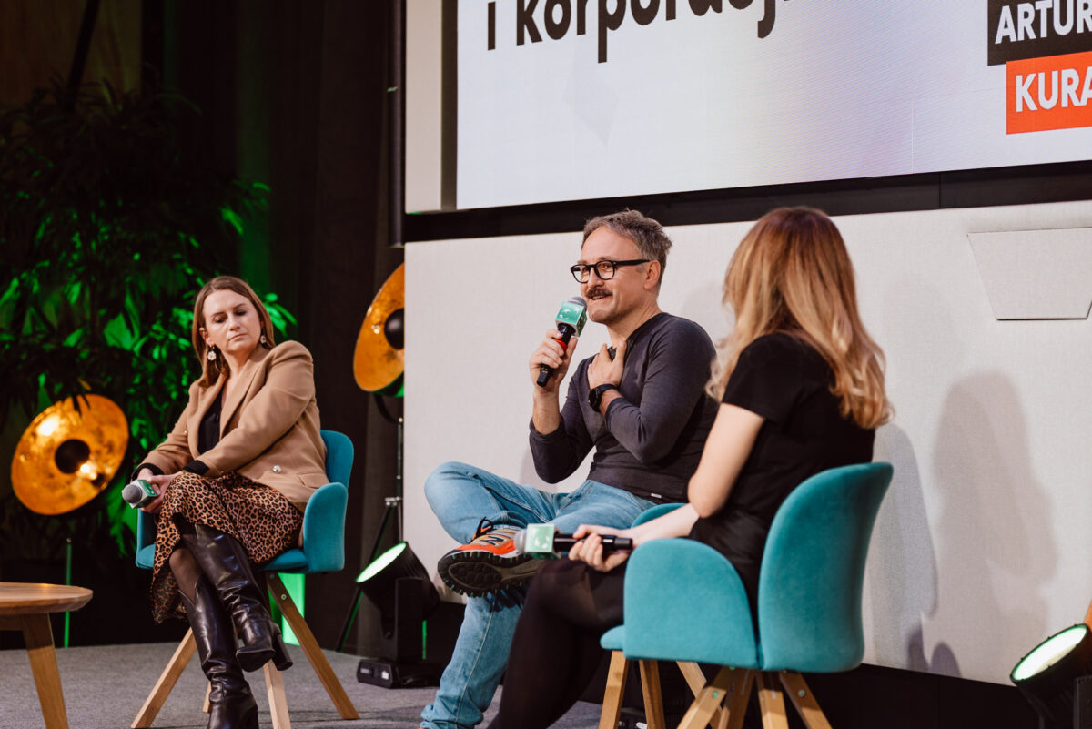 zdjecia eventowe aula polska bnp paribas warszawa 091 Trzy osoby siedzą na scenie podczas dyskusji panelowej. Mężczyzna w środku trzyma mikrofon i mówi, podczas gdy dwie kobiety słuchają. W tle widoczny jest duży ekran i światła sceniczne.