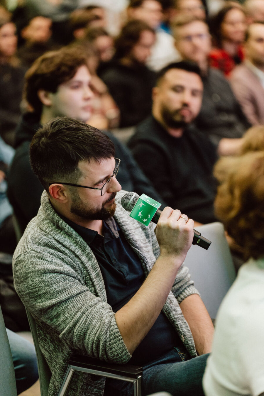 zdjecia eventowe aula polska bnp paribas warszawa 092 Mężczyzna w okularach i z brodą mówi do mikrofonu podczas wydarzenia w pomieszczeniu, otoczony przez innych siedzących uczestników, którzy uważnie słuchają.