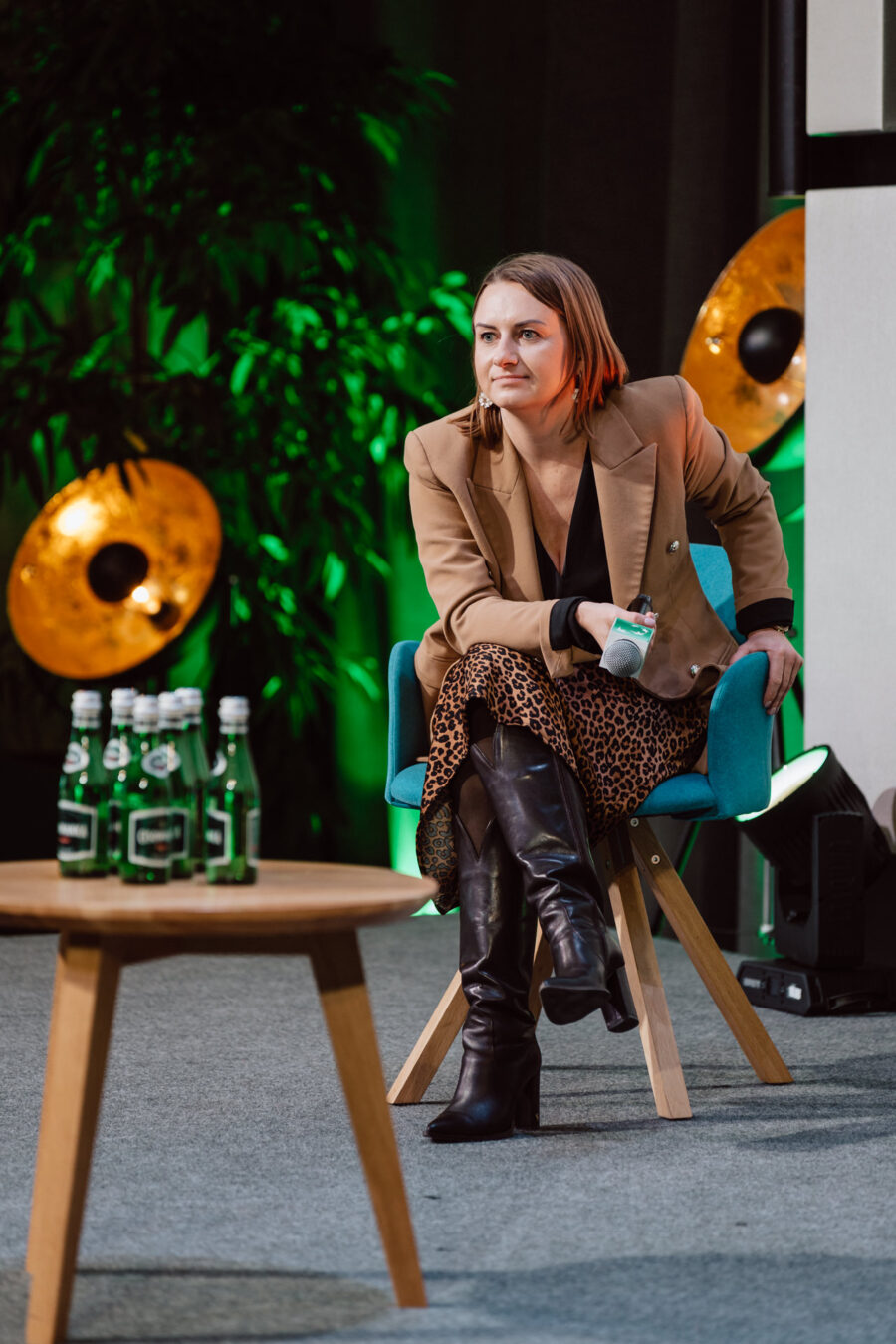 zdjecia eventowe aula polska bnp paribas warszawa 093 Kobieta z krótkimi włosami, ubrana w brązową marynarkę, spódnicę w panterkę i czarne buty, siedzi na niebieskim krześle i trzyma mikrofon. Cztery szklane butelki stoją na drewnianym stole na pierwszym planie. W tle widać zielone oświetlenie.