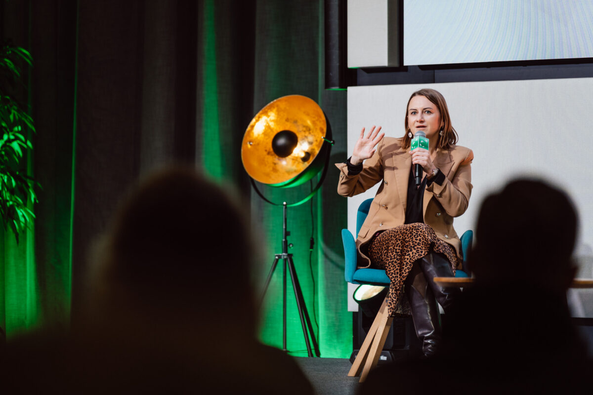 zdjecia eventowe aula polska bnp paribas warszawa 095 Kobieta siedzi na krześle, mówi do mikrofonu i gestykuluje ręką. Ma na sobie brązową marynarkę i spódnicę w lamparcie cętki. Na pierwszym planie widoczna jest publiczność, a w tle znajduje się lampa podłogowa i zielone oświetlenie.
