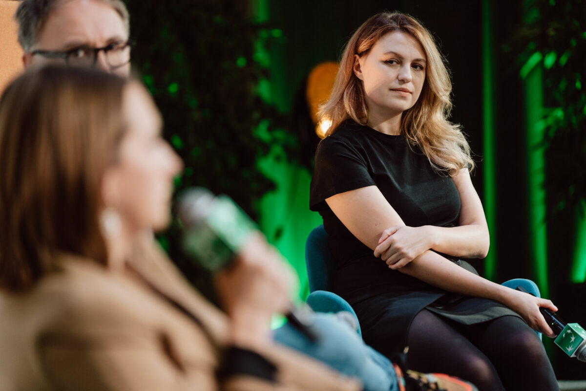 zdjecia eventowe aula polska bnp paribas warszawa 096 Kobieta w czarnej sukience siedzi ze skrzyżowanymi ramionami, uważnie słuchając innej osoby mówiącej do mikrofonu podczas wydarzenia. Tło jest delikatnie oświetlone zielonym światłem.